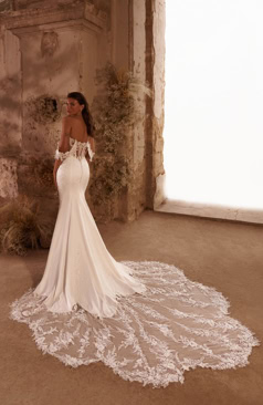 Dieses außergewöhnliche Brautkleid kombiniert verführerische Details mit einem modernen, leichten Look. Das schulterfreie Corsagen-Oberteil aus transparenter Spitze ist kunstvoll bestickt und zaubert mit seinen floralen Applikationen eine sinnliche Silhouette. Die locker fallenden Spitzenärmel betonen Schultern und Dekolleté auf elegante Weise. Der fließende Rock mit hohem Beinschlitz bringt feminine Leichtigkeit und sorgt für einen Hauch Extravaganz. Eine lange Schleppe mit Spitzenkante rundet den Auftritt glamourös ab. Der luftige Schnitt bietet Bewegungsfreiheit und hohen Tragekomfort – perfekt für Bräute, die ihren großen Auftritt modern, selbstbewusst und zugleich romantisch gestalten möchten. Ideal geeignet für Strandhochzeiten, Gartenfeste oder mediterrane Destination Weddings.