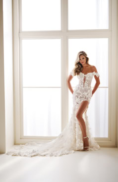 Frau in einem weißen, schulterfreien Brautkleid mit Spitze steht vor einem großen Fenster.